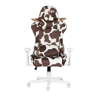 Front view of the cow gaming chair in brown in a white room with grey floor. This cow gaming chair features a white base, with white and brown cow print. The cow chair headrest pillow has brown horns and ears lined with fluffy beige material. The reversible lumbar pillow is shown on the cow print side. This is definitely one of the cutest Techni Sport Gaming chair options and a great option for kawaii gaming setups or as a cute wfh chair. 